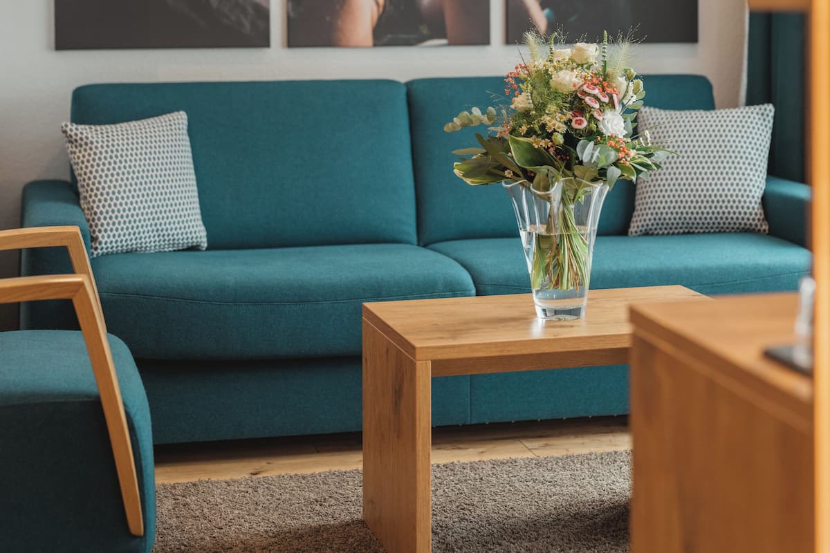 A teal sofa with patterned cushions, a wooden coffee table with a floral arrangement, and a large framed picture of a landscape with a bridge.