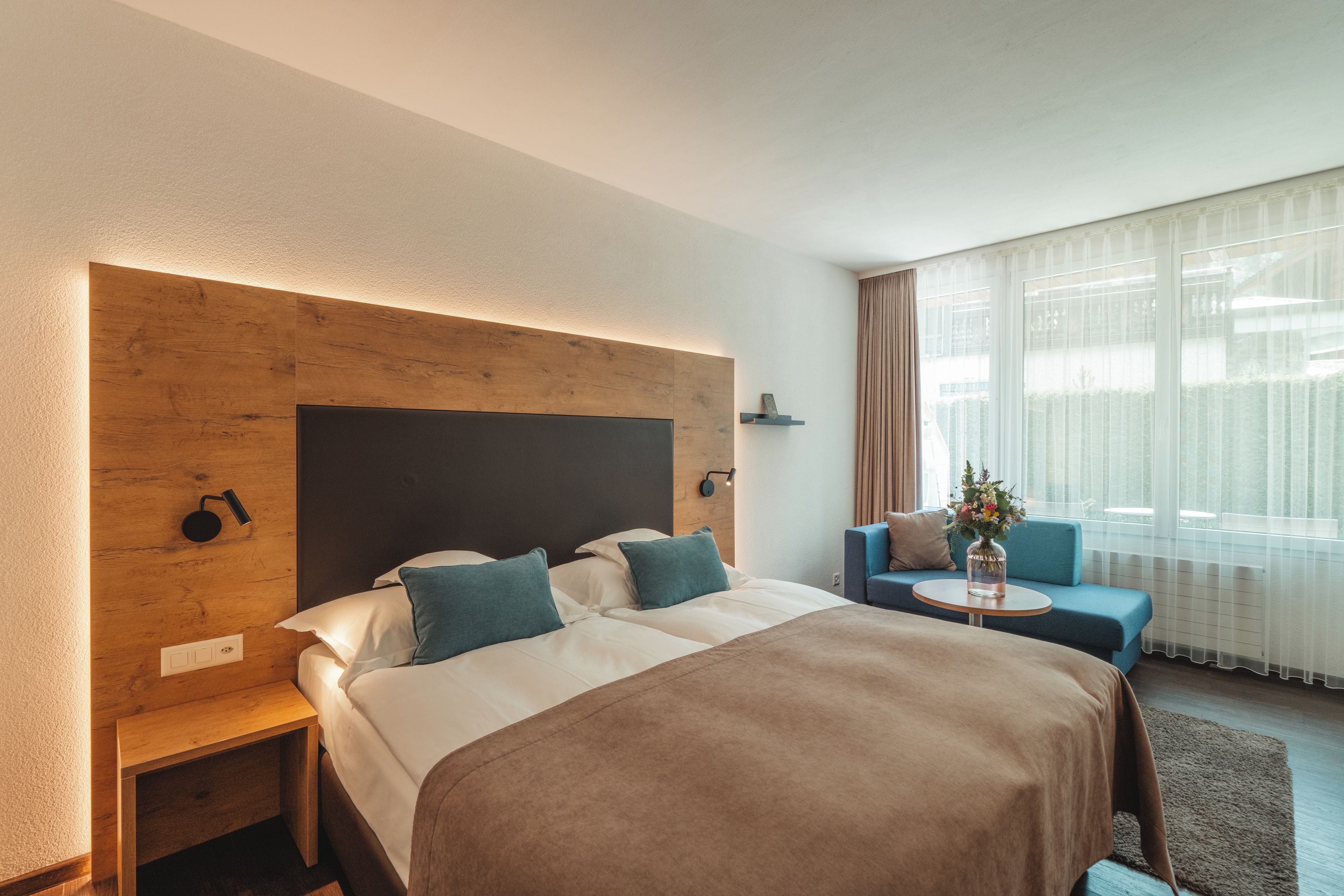 A hotel room with a bed, a nightstand, a blue armchair, a small table with a vase, and a large window with curtains.