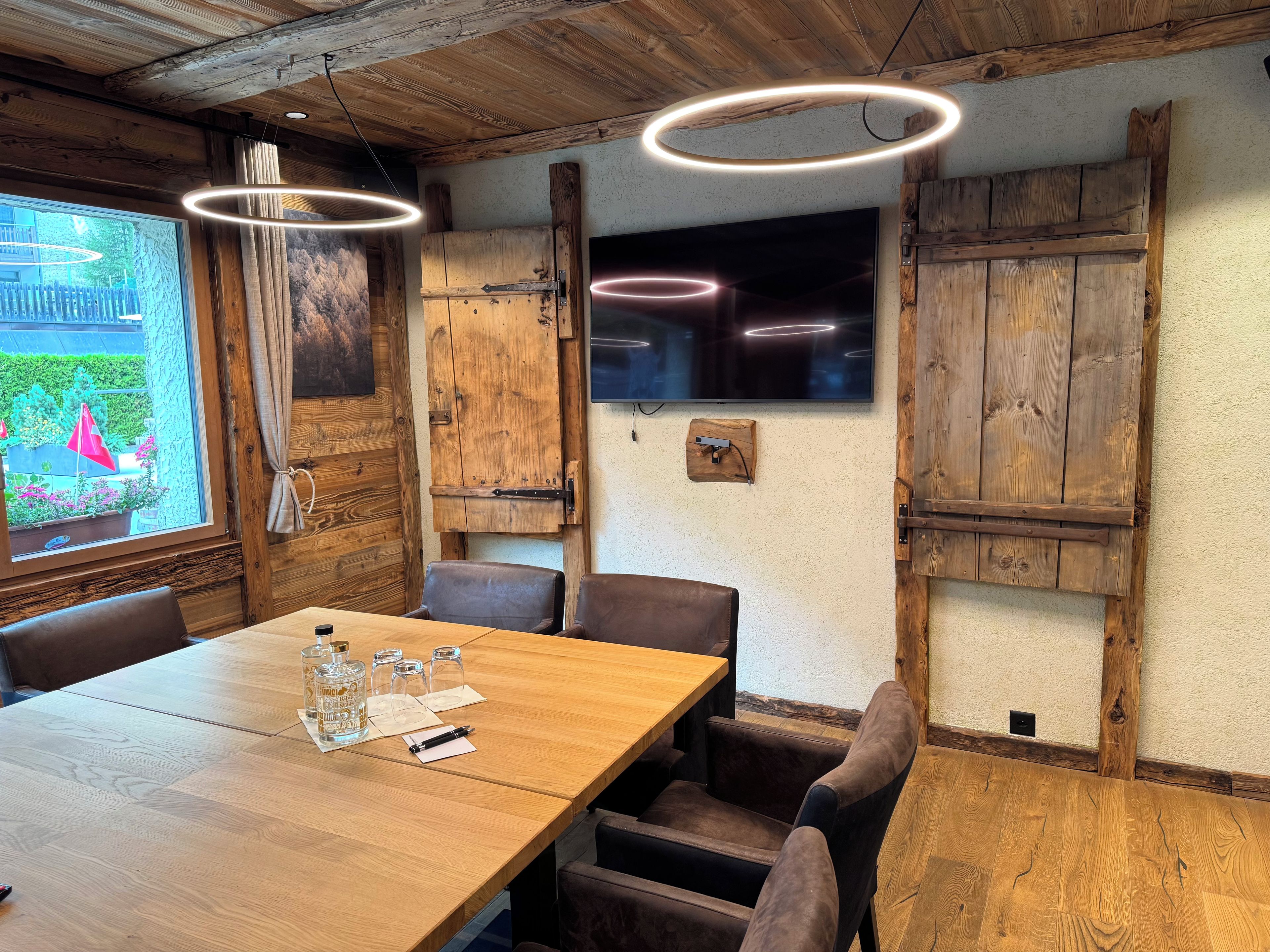 Una mesa de comedor de madera con sillas, un televisor montado en la pared y una ventana con cortinas.