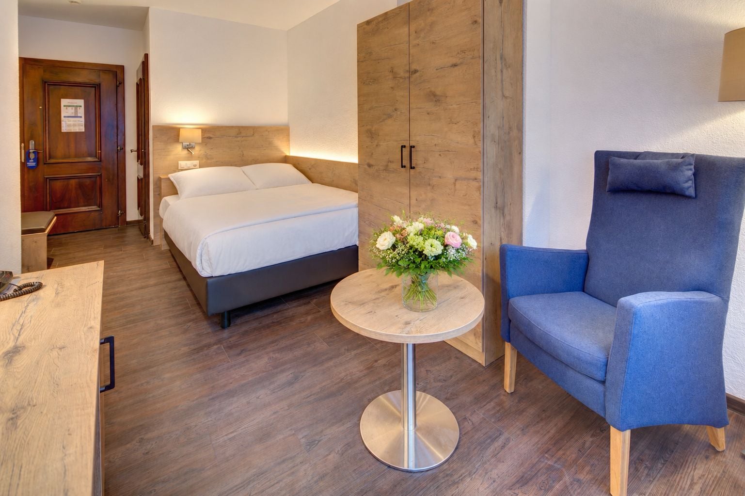A hotel room with a bed, a blue armchair, a small round table with a vase of flowers, and a wooden wardrobe.