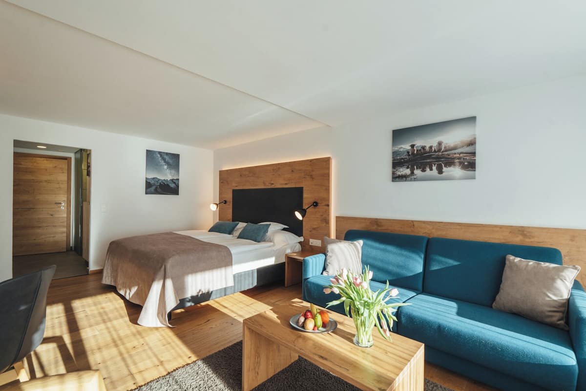 A modern hotel room with a wooden headboard bed, a blue sofa, a wooden coffee table with fruit, and a view of the mountains through the window.