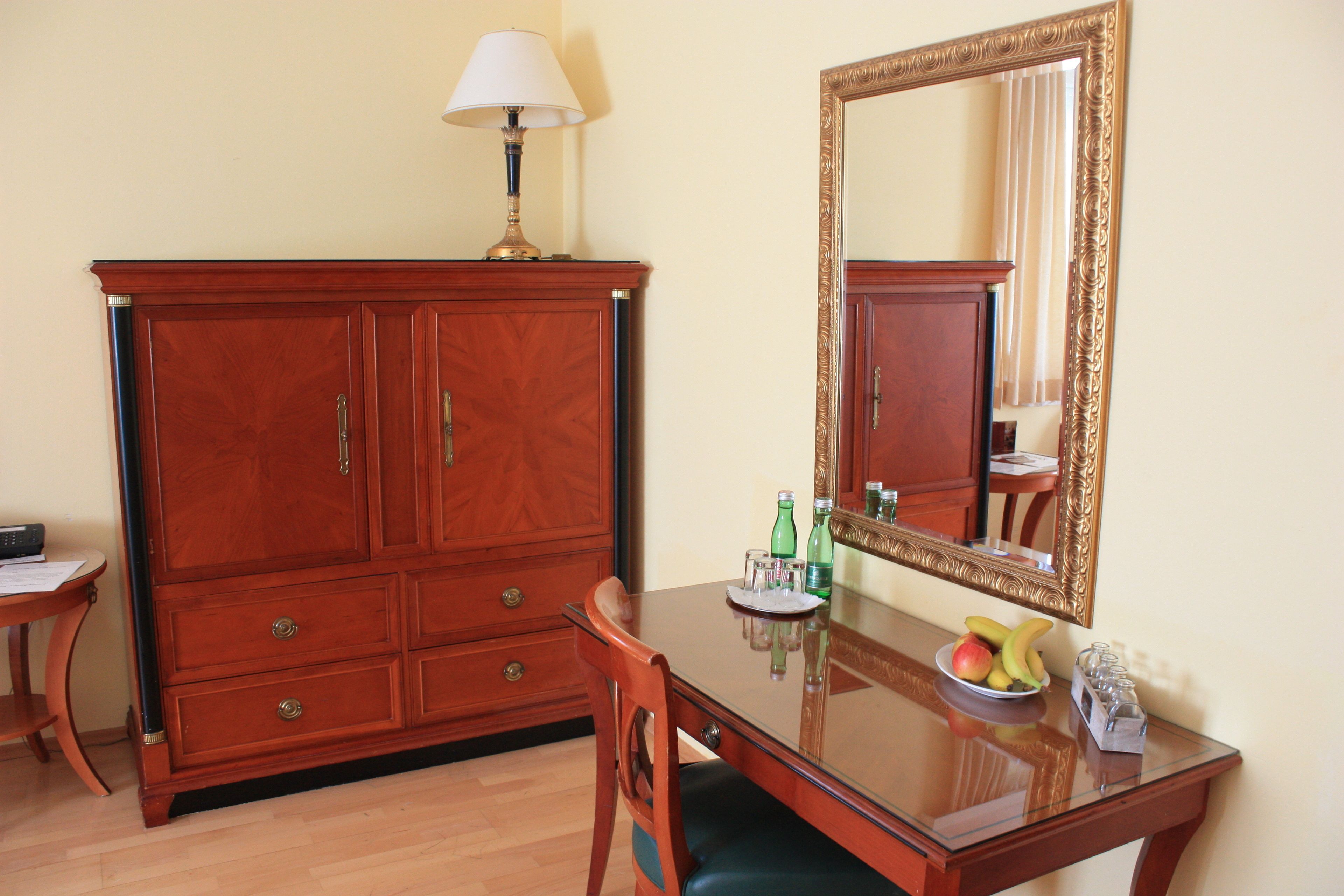 A wooden cabinet with drawers, a table with a glass top, a chair, a mirror with an ornate frame, and a bowl of fruit.