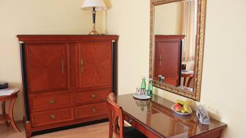 A wooden cabinet with drawers, a table with a glass top, a chair, a mirror with an ornate frame, and a bowl of fruit.