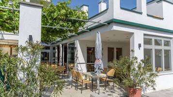A covered outdoor seating area with tables and chairs, surrounded by potted plants and a white building with green trim.
