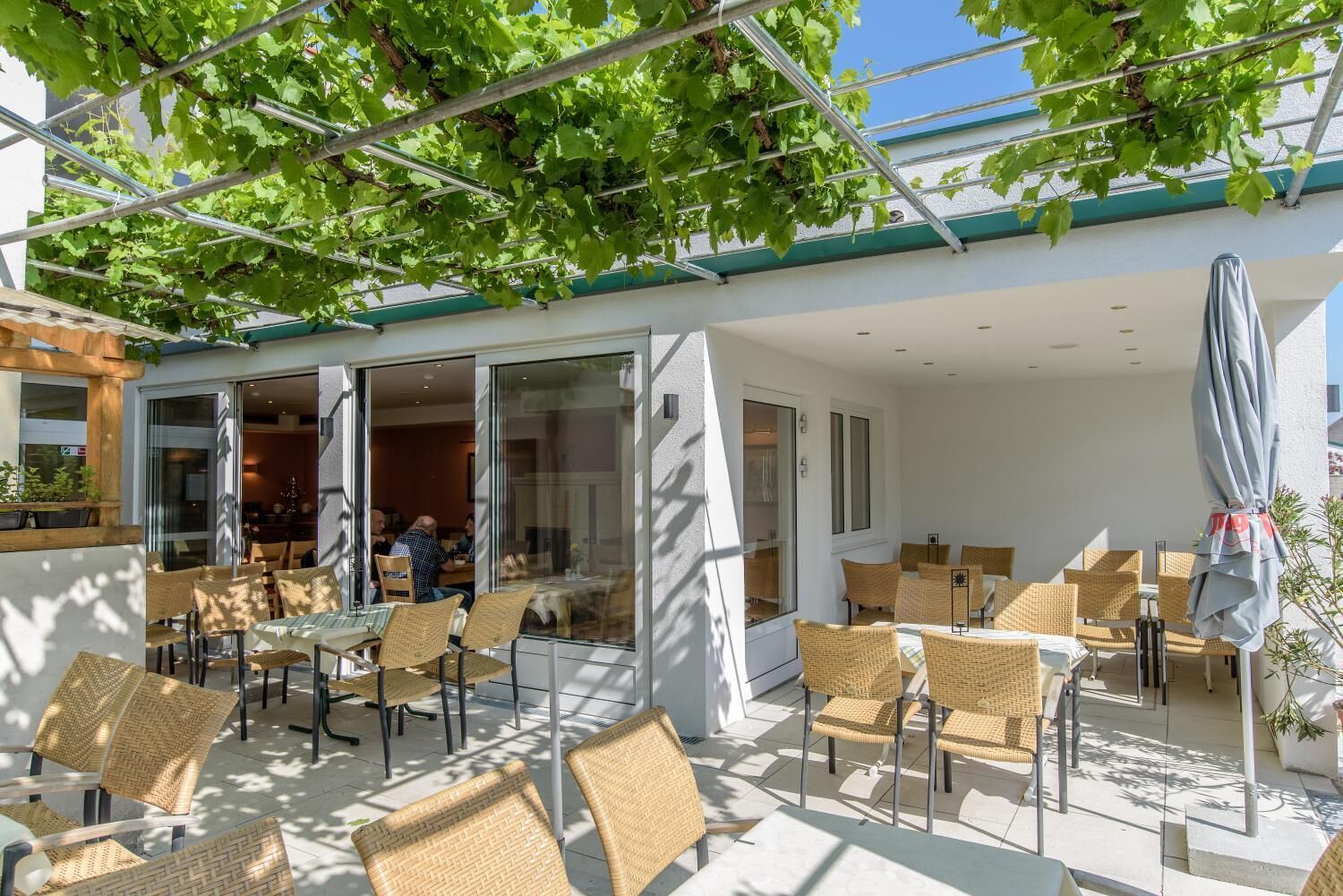 Un patio exterior con mobiliario de mimbre, puertas de cristal que dan a un comedor interior y una pérgola cubierta de enredaderas.