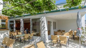 Un patio exterior con mobiliario de mimbre, puertas de cristal que dan a un comedor interior y una pérgola cubierta de enredaderas.