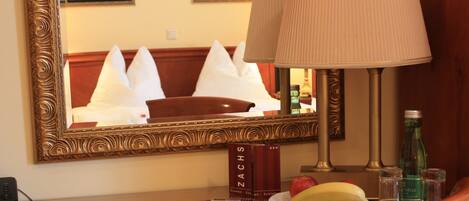 A hotel room with a wooden table, a lamp, a plate with fruit, a bottle, salt and pepper shakers, a framed mirror, and a book titled "ZACHS".