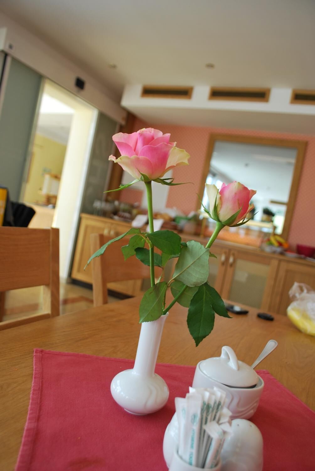 A dining table with a vase of pink roses, a sugar bowl, and a spoon.