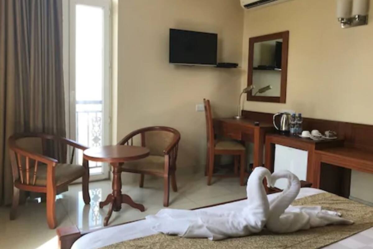 Una habitació d'hotel amb un llit, una taula de nit, dues cadires, un televisor i una tovallola amb forma de cigne sobre el llit.