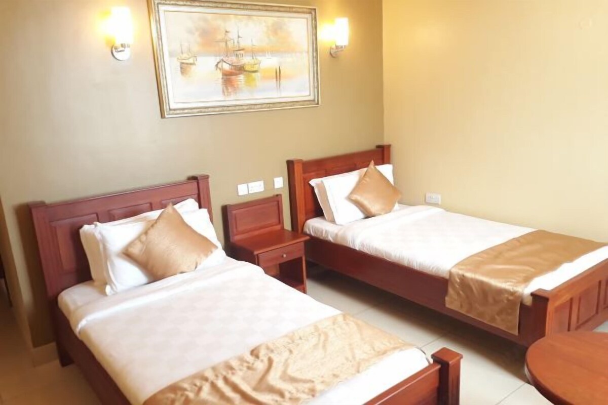 A hotel room with two single beds, wooden headboards, bedside tables, and a framed picture on the wall.