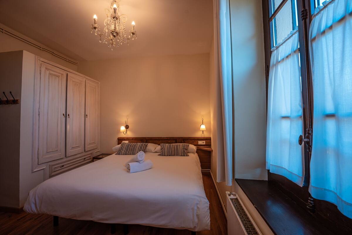 Un lit bien fait, avec une serviette pliée, un lustre et une grande fenêtre avec des rideaux.