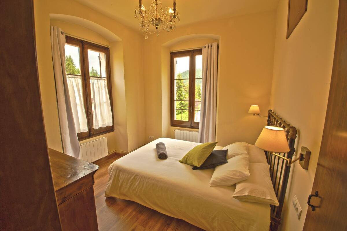 Une chambre à coucher avec un grand lit, deux fenêtres avec des rideaux, un lustre et une lampe de chevet.