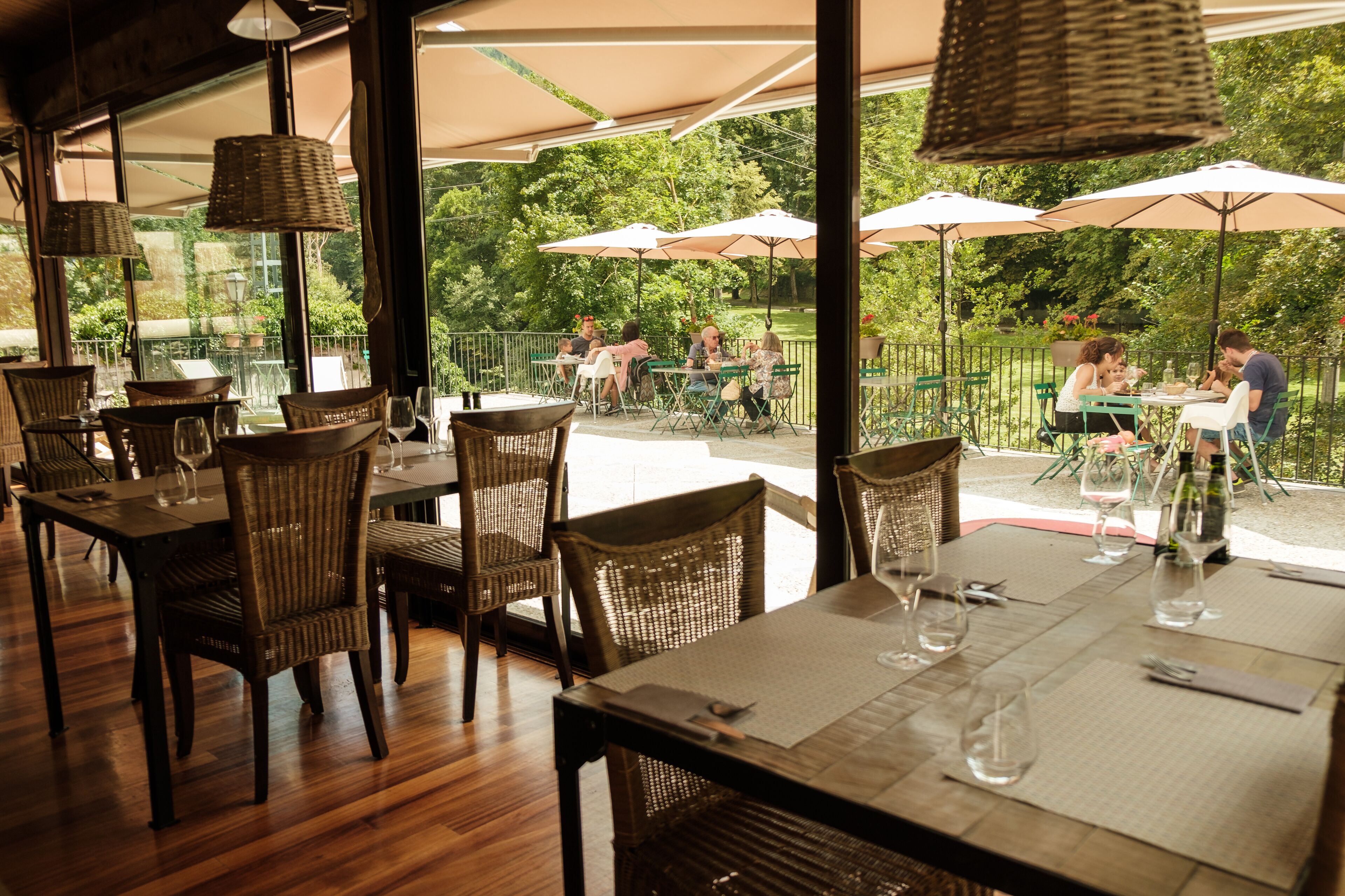 Un espace de restauration intérieur avec des chaises et des tables en osier, de grandes fenêtres et un espace extérieur aménagé avec des parasols.