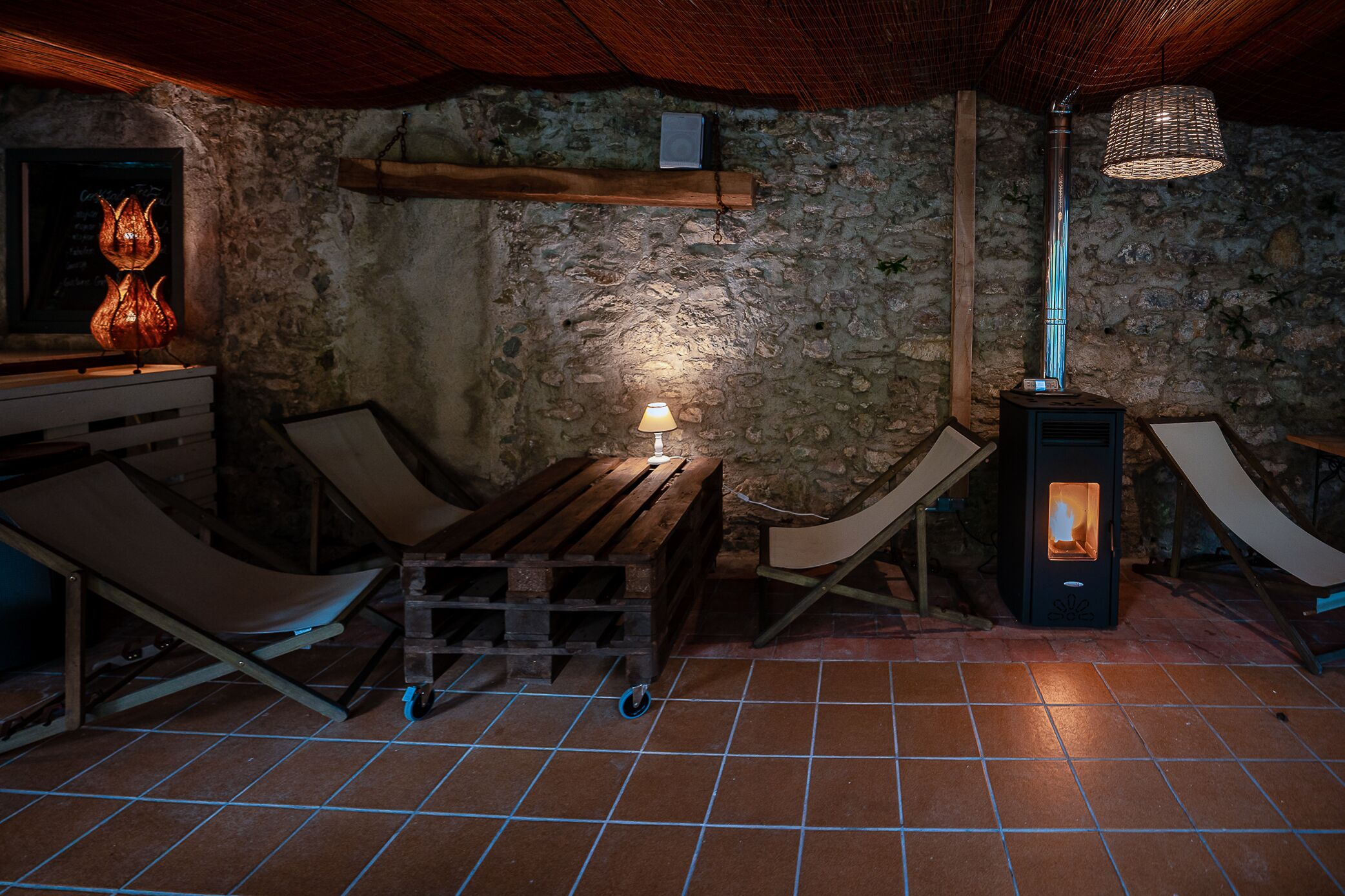 A rustic interior with a stone wall, a wooden bench, a table, a hanging light fixture, and a wood-burning stove.