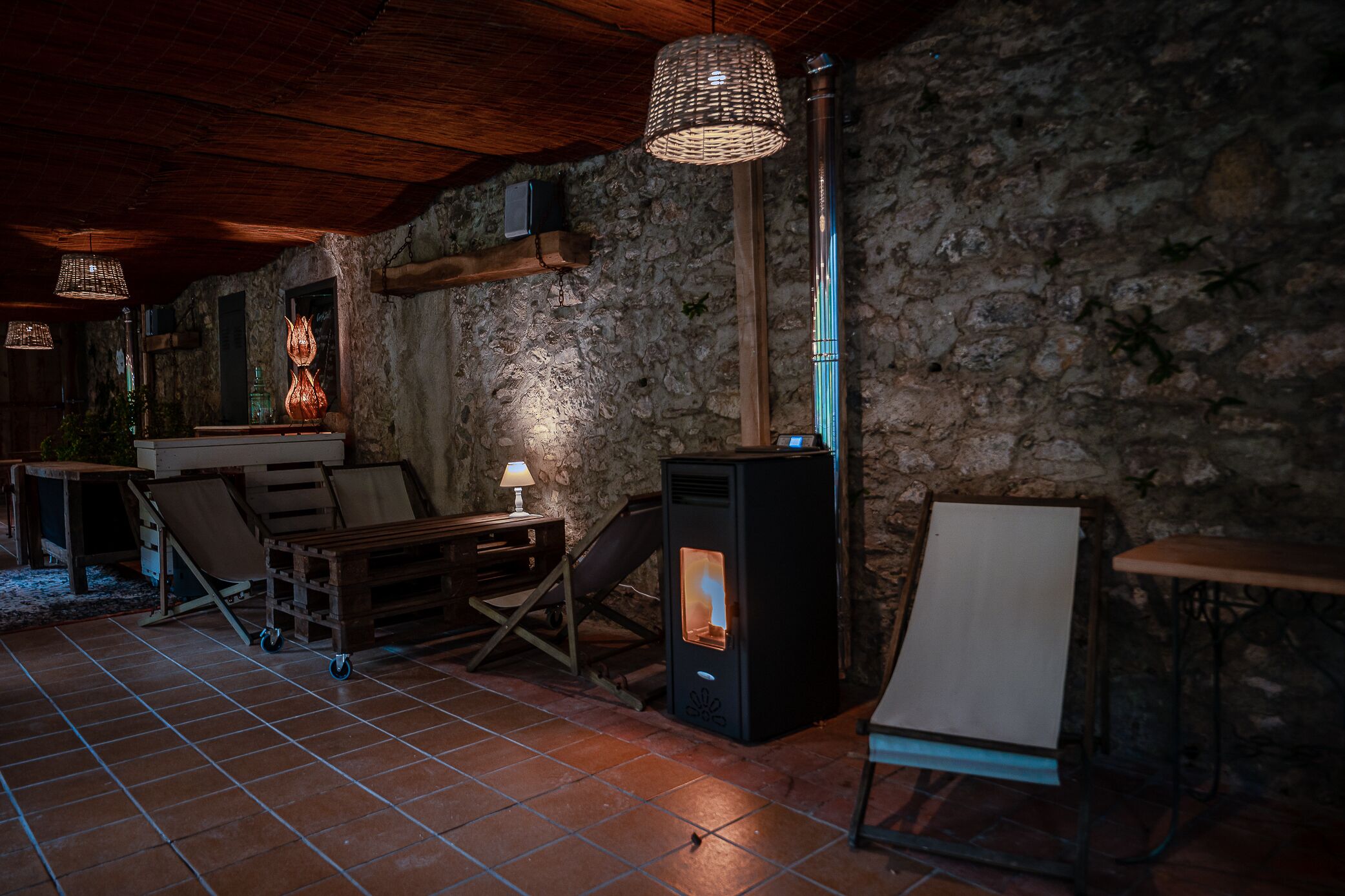 Un interior rústico con pared de piedra, estufa de leña y una mesa de madera con lámpara.