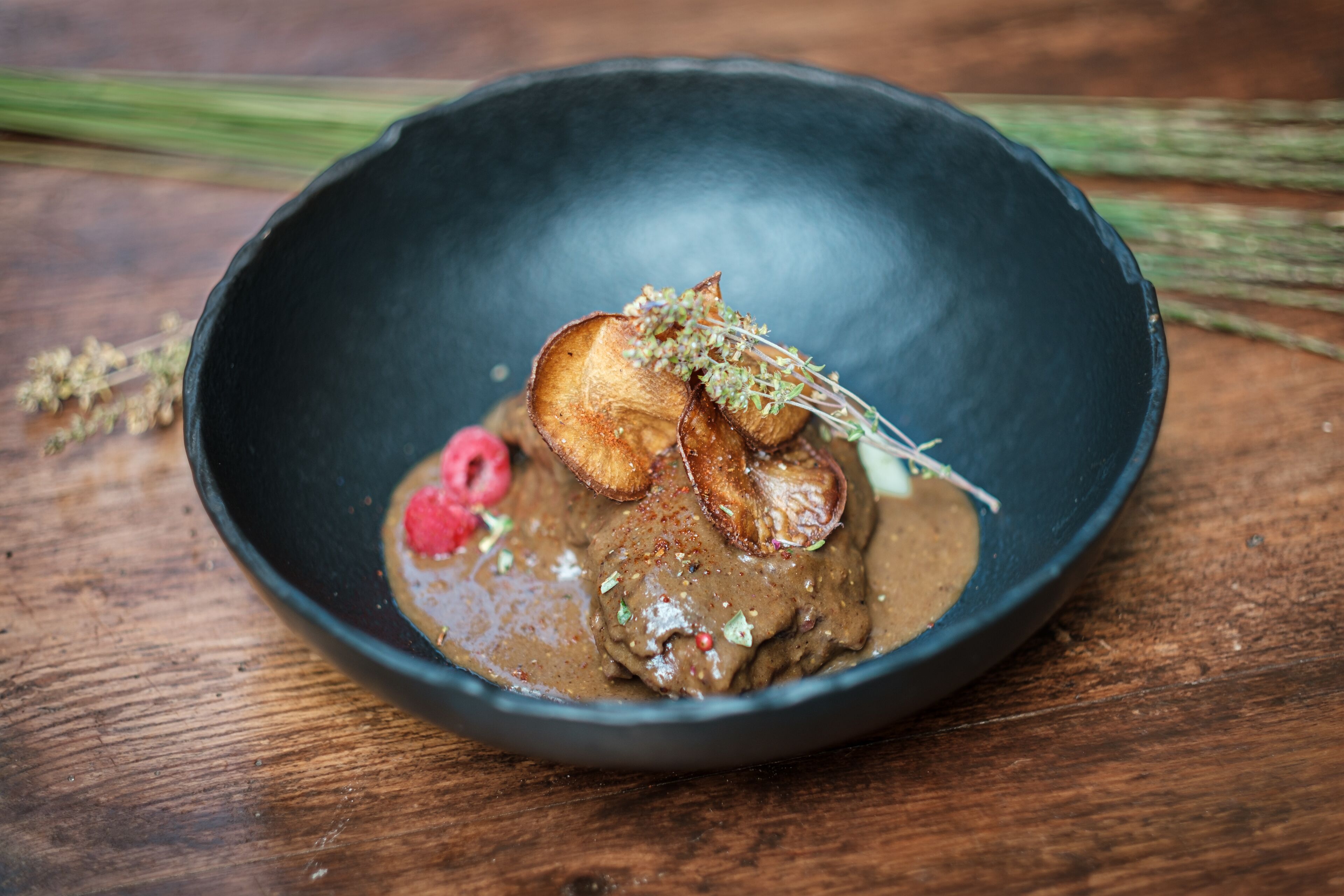A dark bowl with a creamy sauce, mushrooms, and a sprig of herbs on a wooden surface.