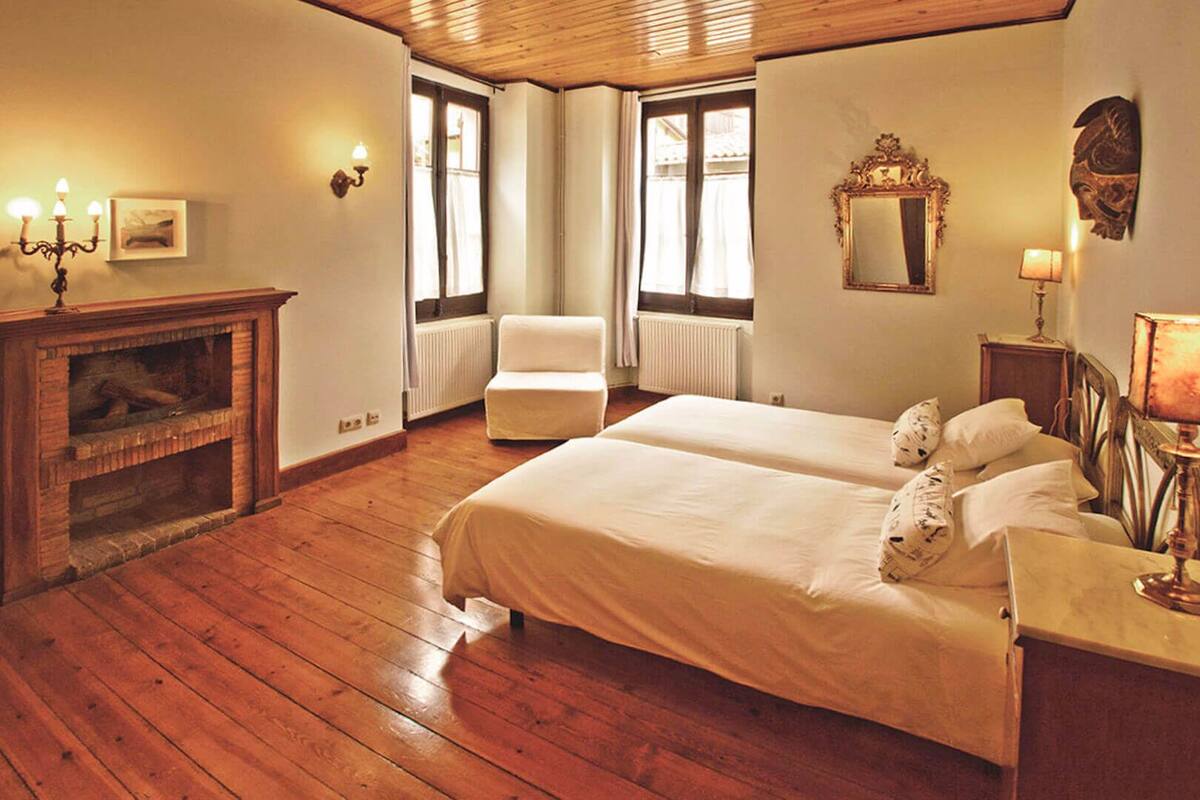 Une chambre avec un plancher en bois, une cheminée, deux lits, une chaise et un miroir.
