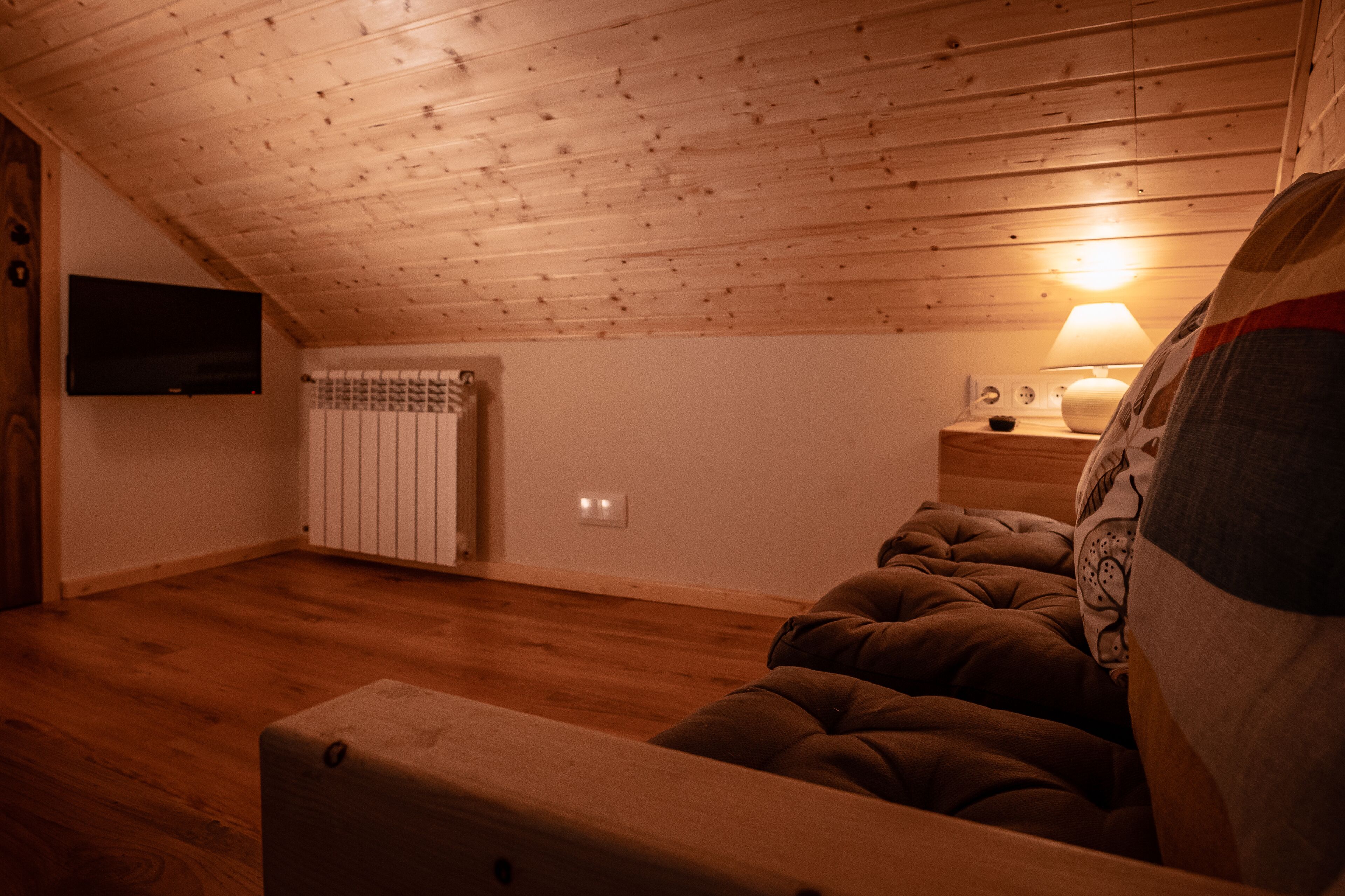 Un dormitorio acogedor en el ático, con techo de madera, una cama, una mesita de noche con una lámpara y un televisor montado en la pared.