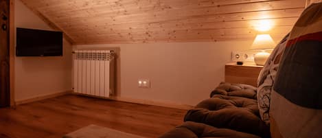 A cozy attic bedroom with wooden ceiling, a bed, a nightstand with a lamp, and a TV mounted on the wall.