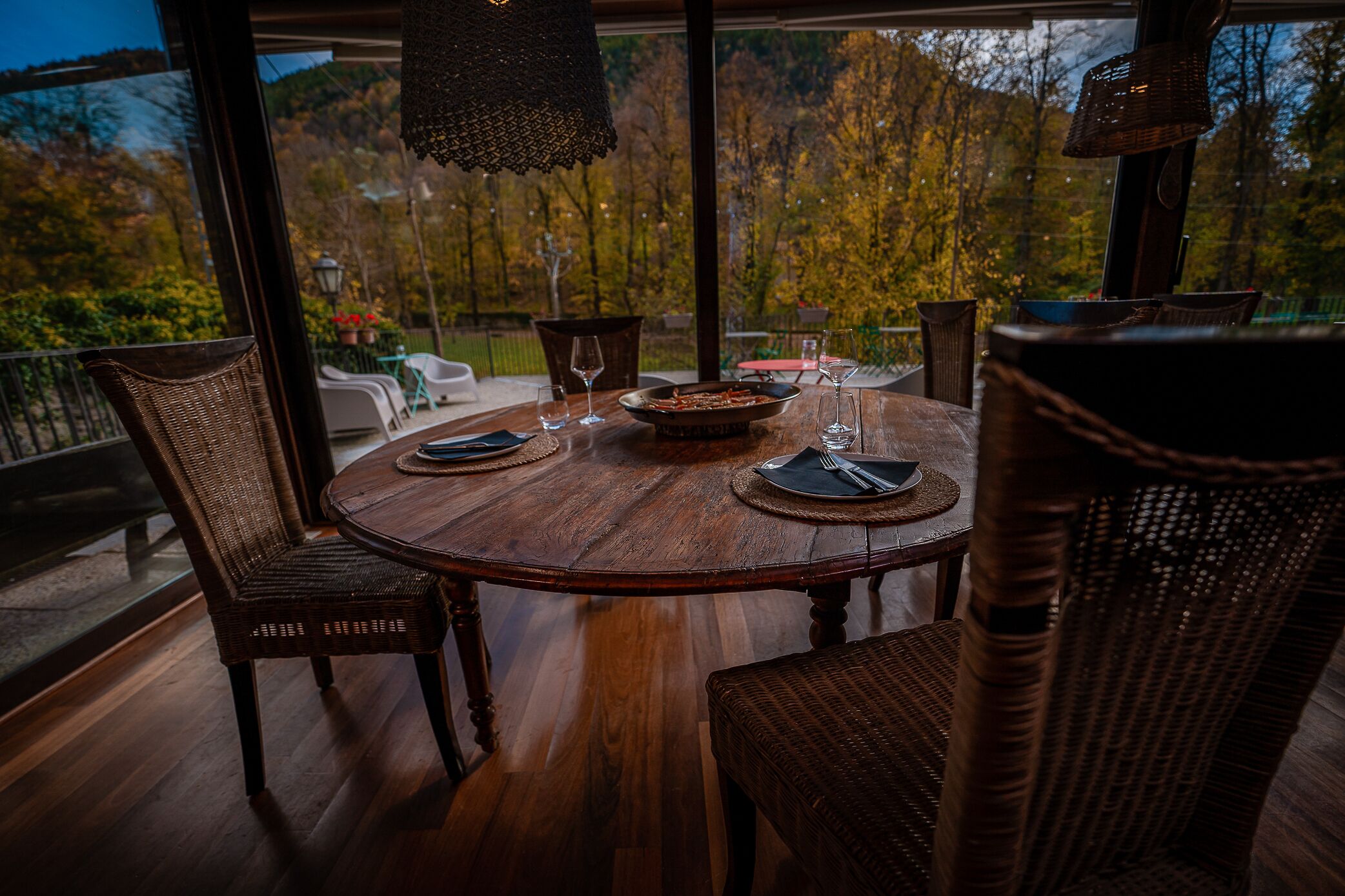 A round wooden dining table with wicker chairs, set for a meal with plates, cutlery, and glasses.