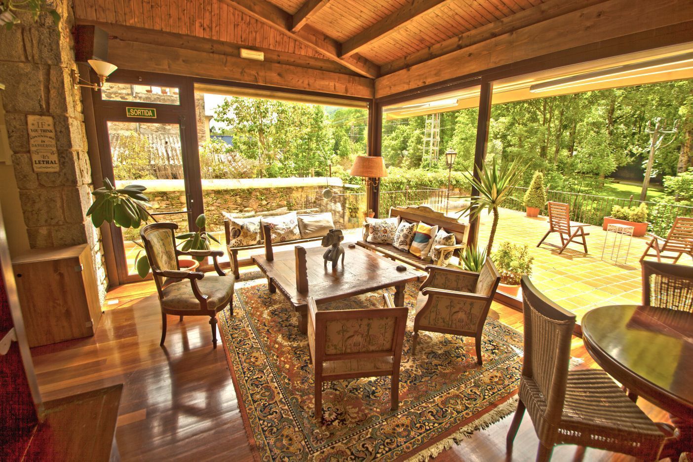 A cozy, well-lit patio area with a wooden ceiling, a dining table surrounded by chairs, and a view of lush greenery outside.