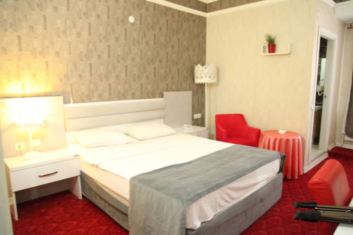 A hotel room with a bed, nightstand, lamp, red chair, small table, and a mirror.