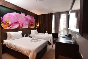 Habitación de hotel con dos camas, un escritorio, una silla, un cuadro grande de una orquídea y vistas de edificios por la ventana.
