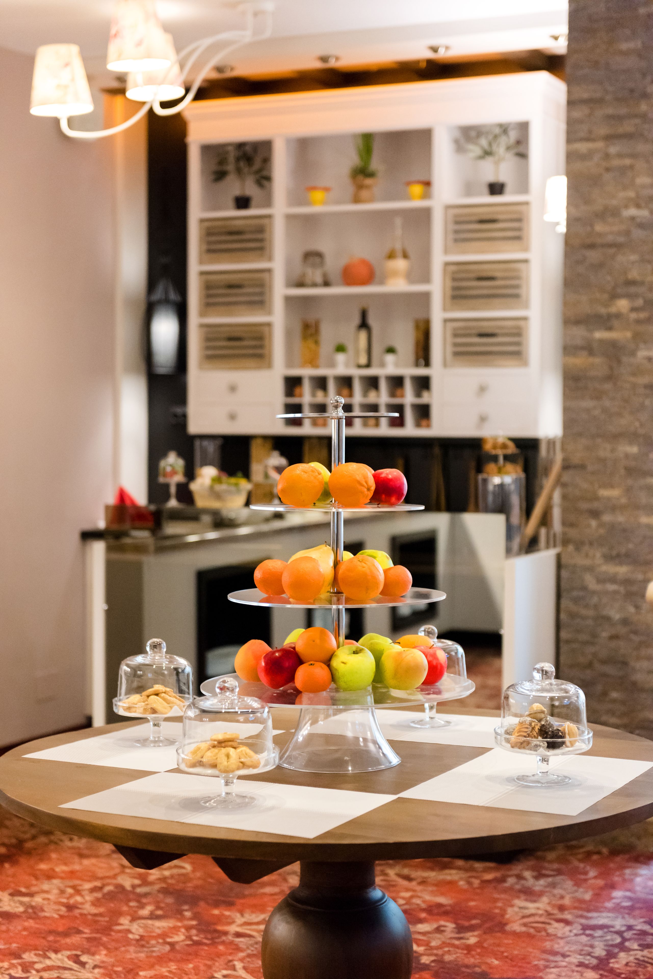 A three-tiered fruit stand with apples, oranges, and pears, and small dessert dishes on a table in a room with a kitchen in the background.