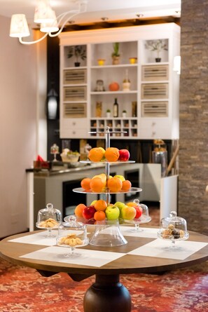 Un puesto de frutas de tres niveles con manzanas, naranjas y peras, y pequeños recipientes de postre sobre una mesa en una habitación con una cocina al fondo.