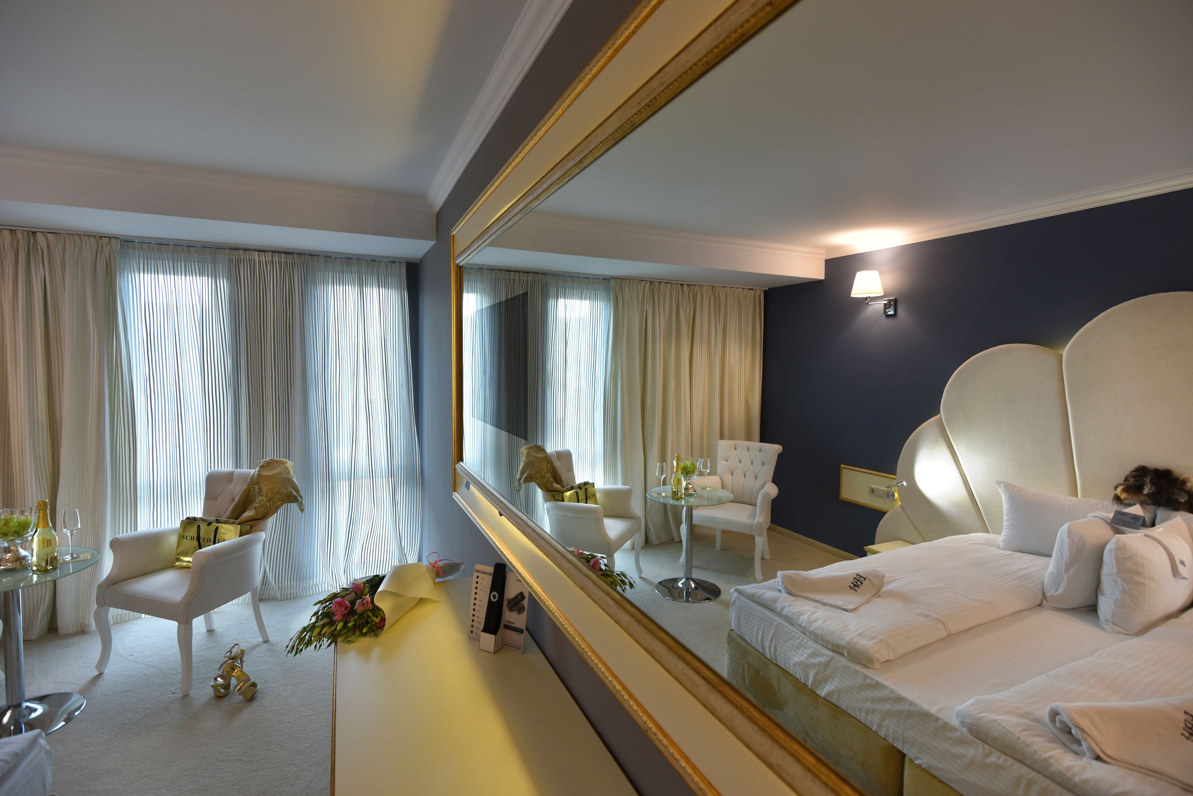 A hotel room with a large mirror, a bed with white linens, a round table with a chair, and a small table with a bouquet of flowers and a bottle of champagne.