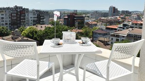 Un balcón con mesa y sillas, con vista a un paisaje urbano con edificios y árboles.