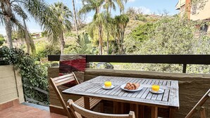Una mesa de madera con dos platos, uno con un croissant y dos vasos de jugo de naranja, en un balcón con vista a follaje y un edificio al fondo.