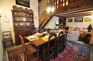 Un comedor con una mesa de madera dispuesta para cuatro personas, rodeada de sillas. A la izquierda, un aparador de madera con varios platos es visible. Una escalera conduce a un nivel superior.