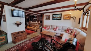 Ein gemütliches Wohnzimmer mit Sofa, Hocker und Couchtisch. Ein Bücherregal voller Bücher und ein flachbildfernseher an der Wand sind ebenfalls vorhanden.