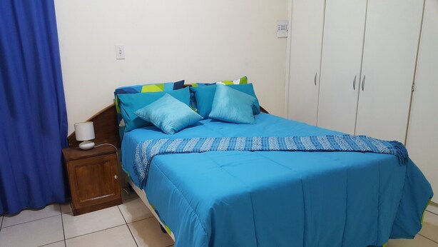 A bedroom with a bed covered in blue bedding, a wooden nightstand with a lamp, and a wardrobe. - Indigo Guesthouse (Pinetown)