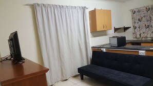 A small, compact room with a wooden desk, a black chair, a small kitchenette, and a curtain. - Indigo Guesthouse (Pinetown)