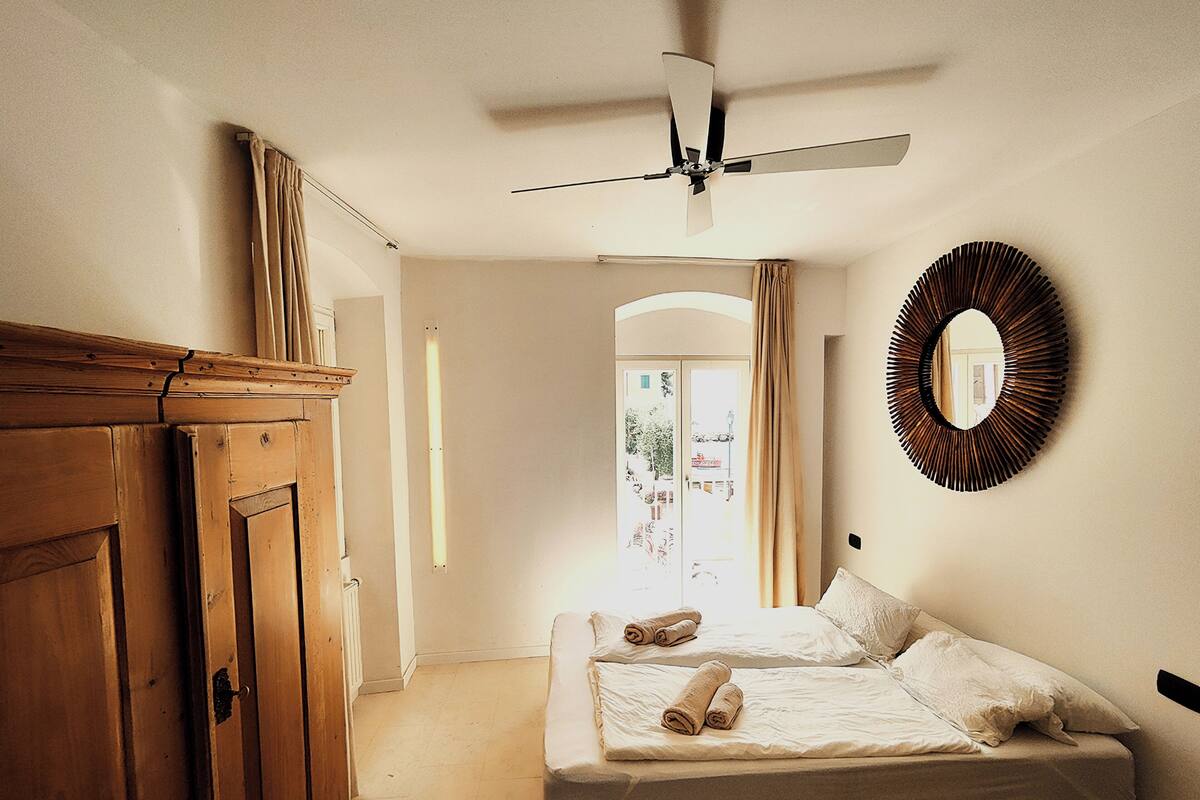 A bedroom with a bed, a ceiling fan, a wooden wardrobe, and a mirror on the wall.
