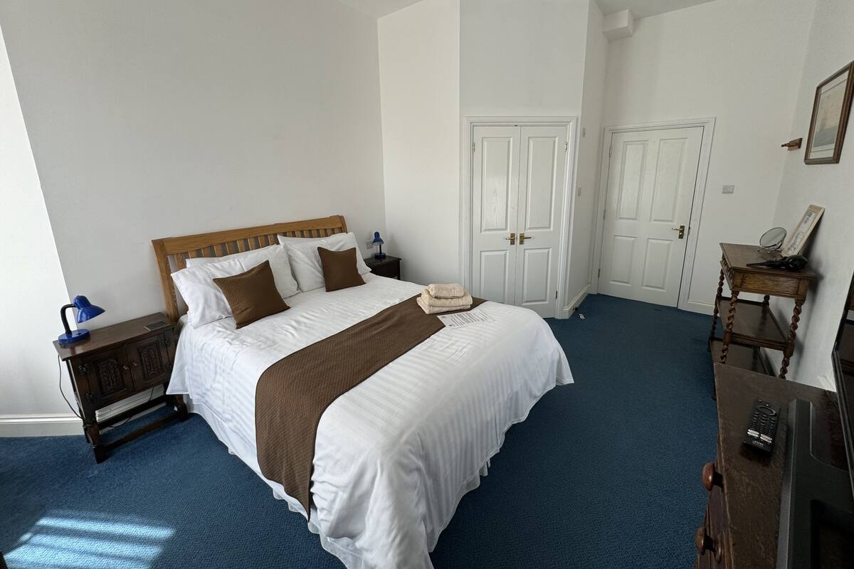 A neatly arranged hotel room with a large bed, a bedside table with a lamp, a dresser, and a door leading to another room.