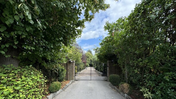 A paved driveway leading to a gated entrance surrounded by dense foliage. - Hookwood Lodge (Horley)