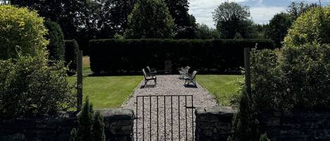 A stone pathway leading to a seating area with two chairs, surrounded by lush greenery and a clear sky.