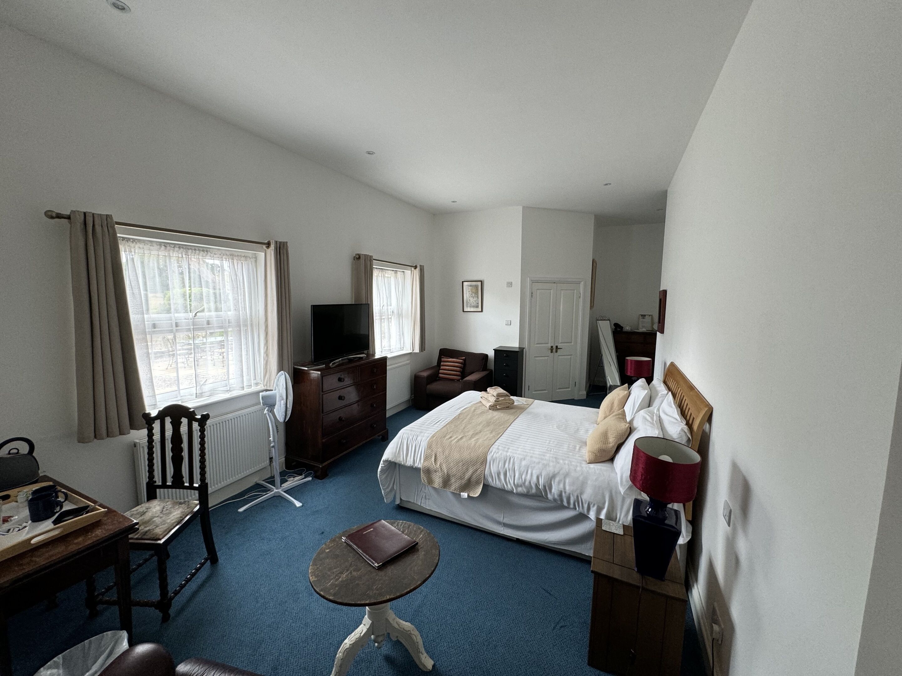 Habitación de hotel con cama, escritorio y silla, una mesita, cómoda, televisor y ventana con cortinas.