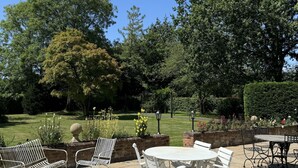 An outdoor patio with white metal furniture, including chairs and a table, surrounded by a brick wall and a well-maintained garden with various plants and flowers. - Hookwood Lodge (Horley)
