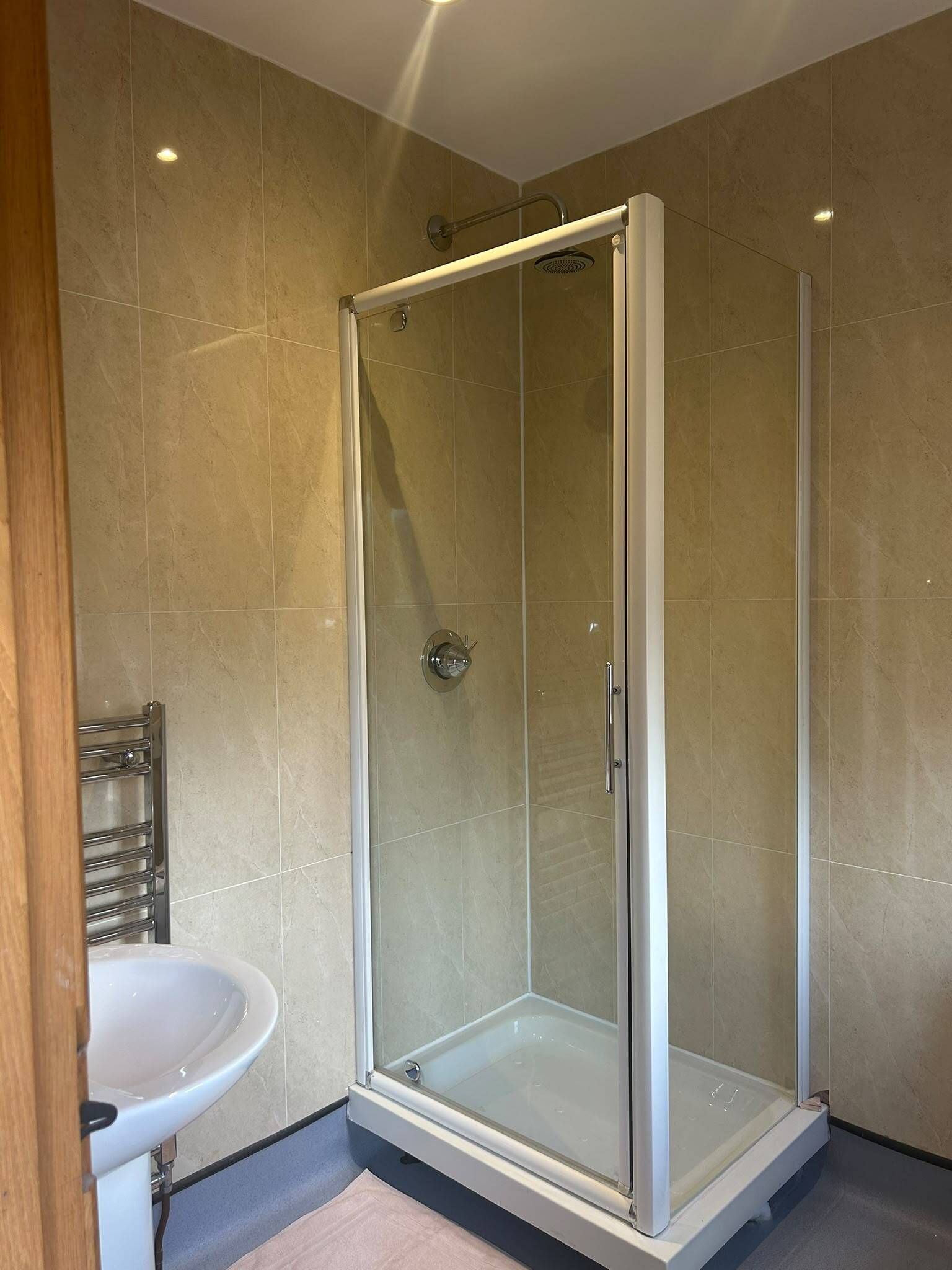 Et brusebad med glasvægge og hvid ramme, en hvid vask og vægge med beige fliser.