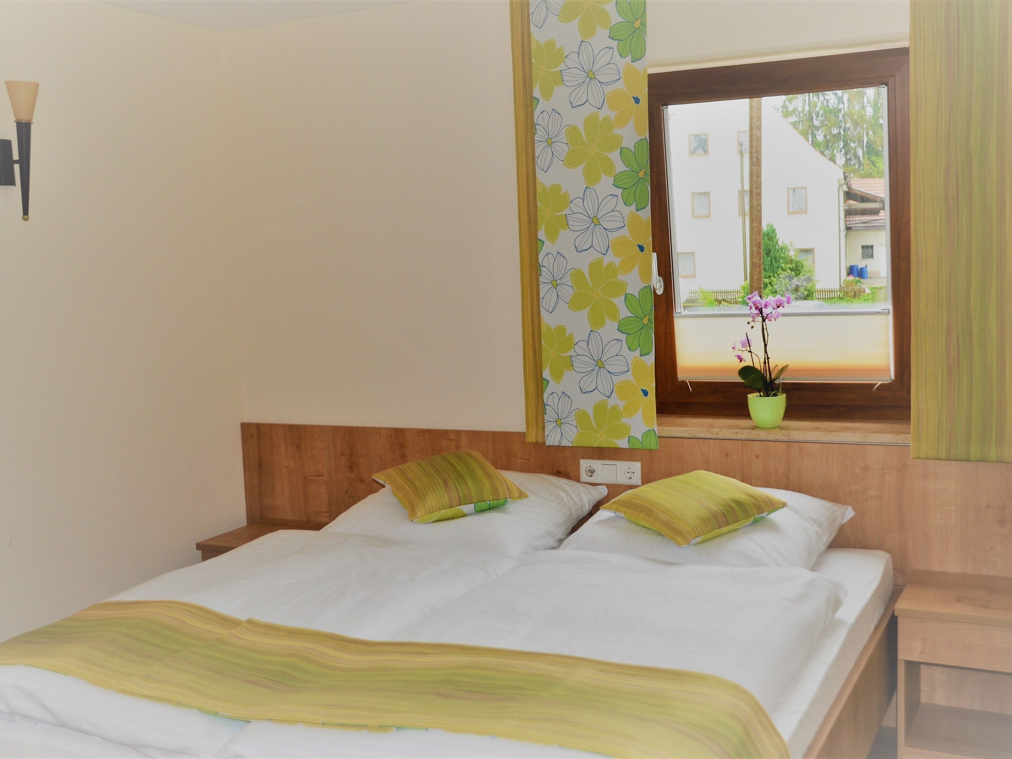 A bedroom with a bed, pillows, a window with a view of buildings, a wall-mounted lamp, and a potted plant on the windowsill.