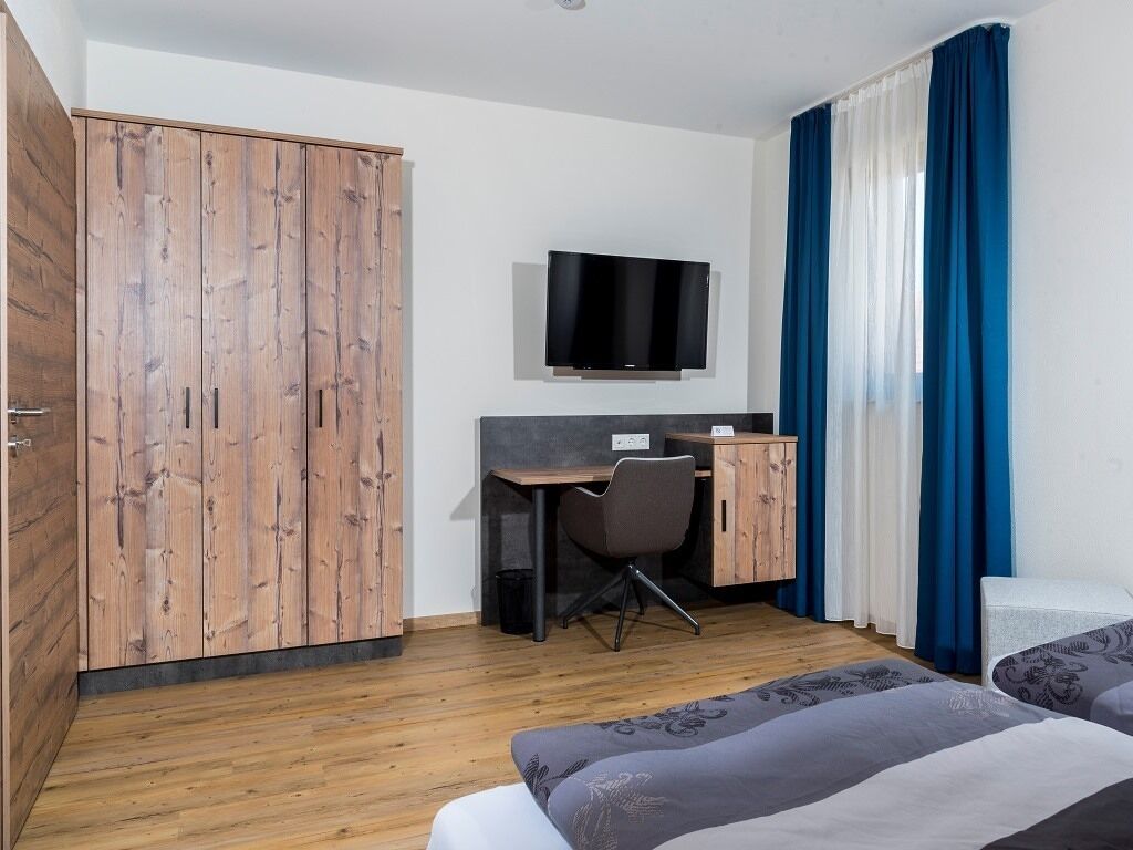 Une chambre d’hôtel avec une armoire en bois, un bureau avec une chaise, un téléviseur fixé au mur et un lit avec une literie à motifs.