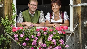 Deux personnes, vêtues de costumes traditionnels, sont assises à une table en bois devant laquelle se trouvent des fleurs roses.