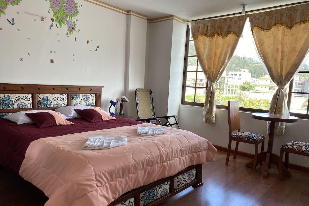 A hotel room with a large bed, a desk with a chair, a mirror, and a window with curtains.