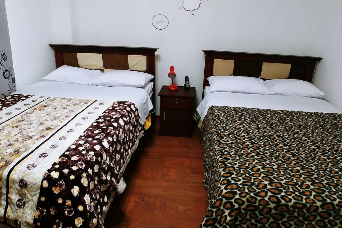 A hotel room with two beds, each with a different patterned bedspread. There is a nightstand with a lamp and a phone between the beds. The walls are plain with a decorative wall hanging above the beds.