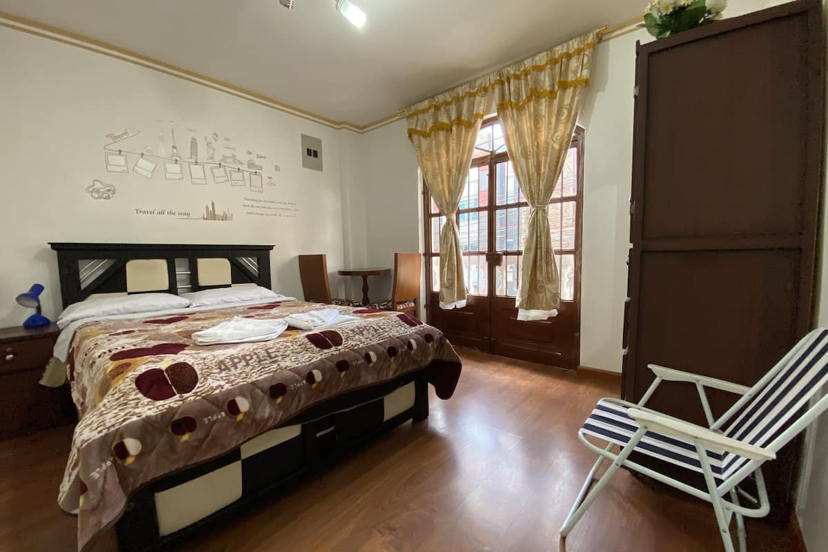 A hotel room with a bed, a chair, a desk, a wall with a cityscape mural, and a window with curtains.
