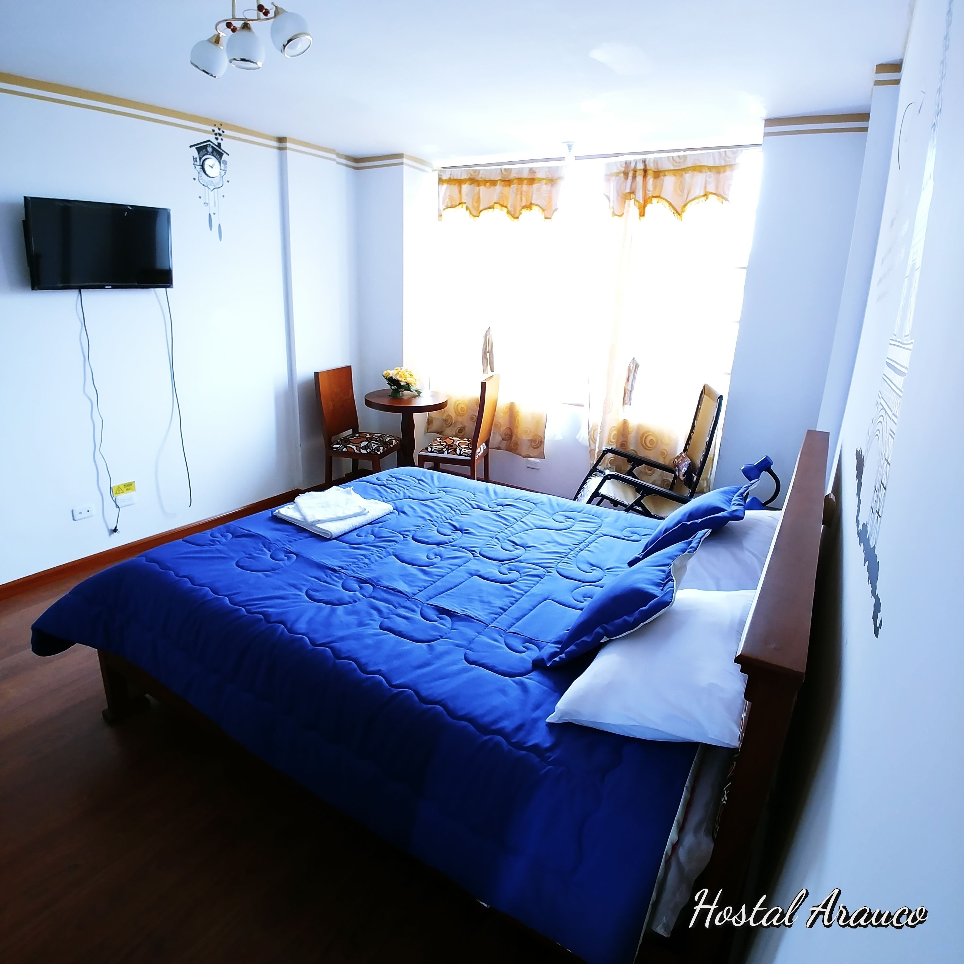 A hotel room with a large bed, a flat-screen TV mounted on the wall, a small table with chairs, and a window with curtains.